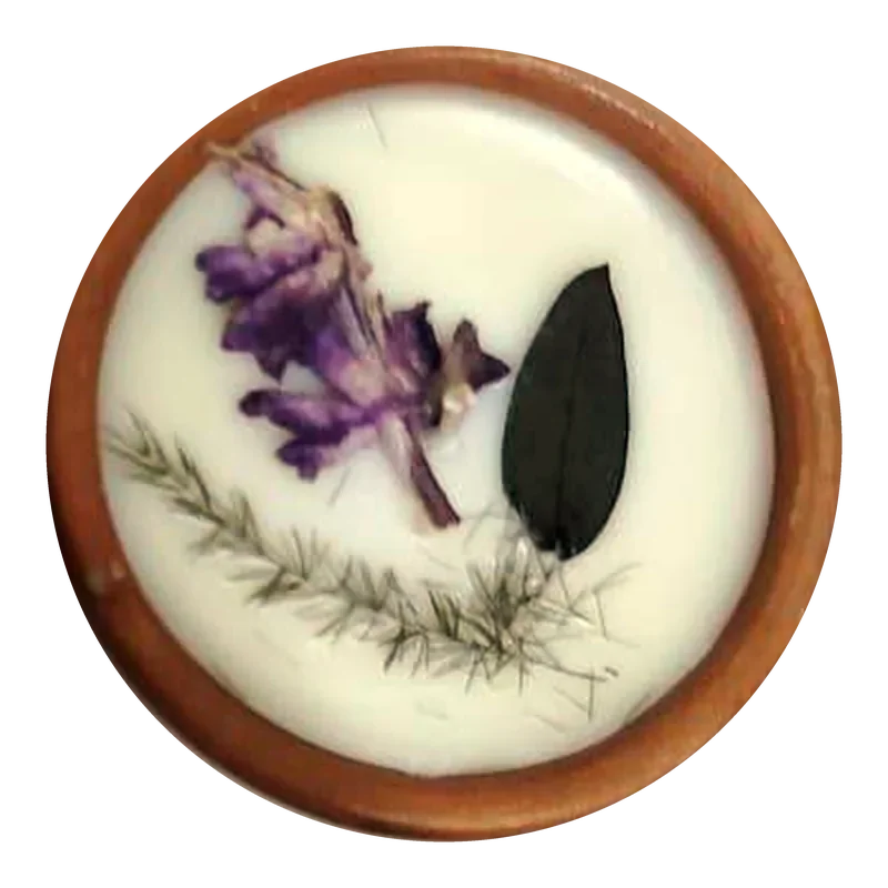 Close-up of a terracotta diya filled with white soy wax, decorated with a pressed lavender-violet flower, a single deep-green leaf and a fern sprig.