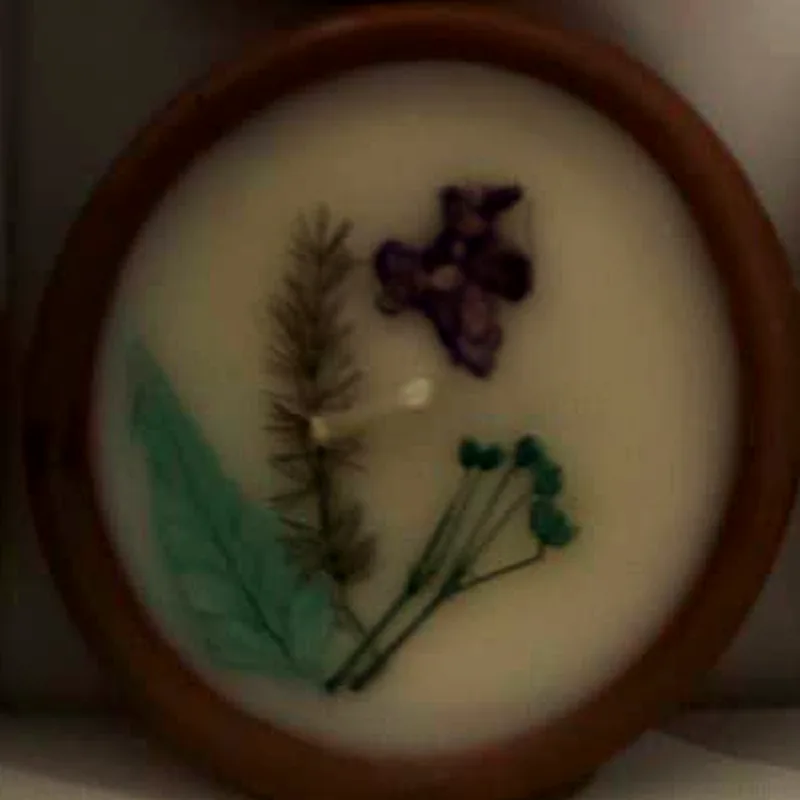 A single diya with a mint-green leaf, a fern sprig and a small violet flower pressed into white wax.