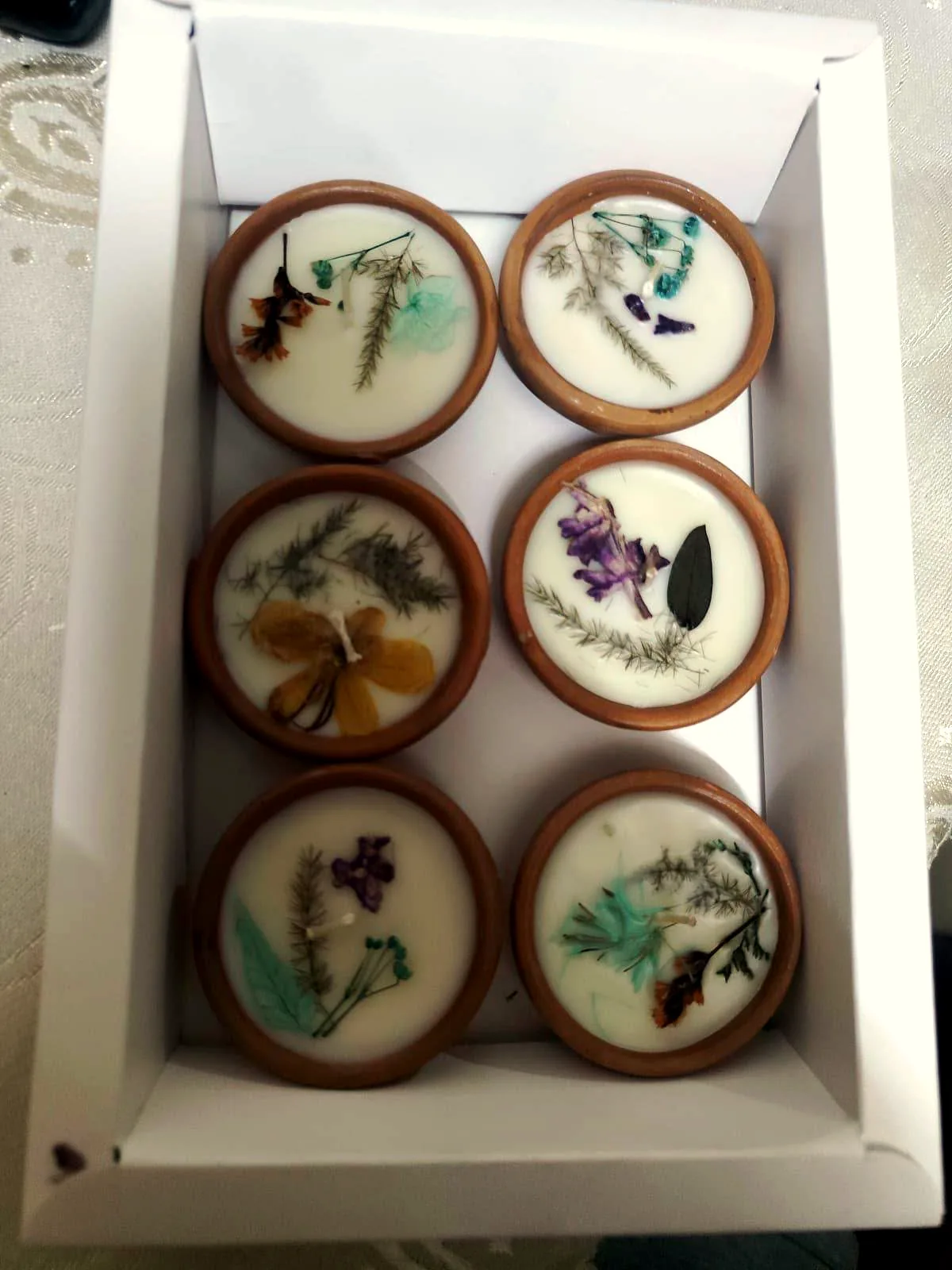 An open white gift box holding six terracotta diyas in two rows. Each diya is filled with white wax and decorated with different pressed dried flowers and fern sprigs in orange, purple, turquoise and gold.